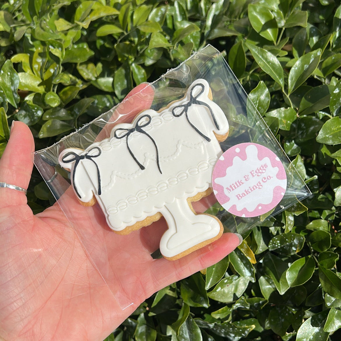 Hand holding a cookie shaped like a cake with a black coloured bows against a green leafy background.