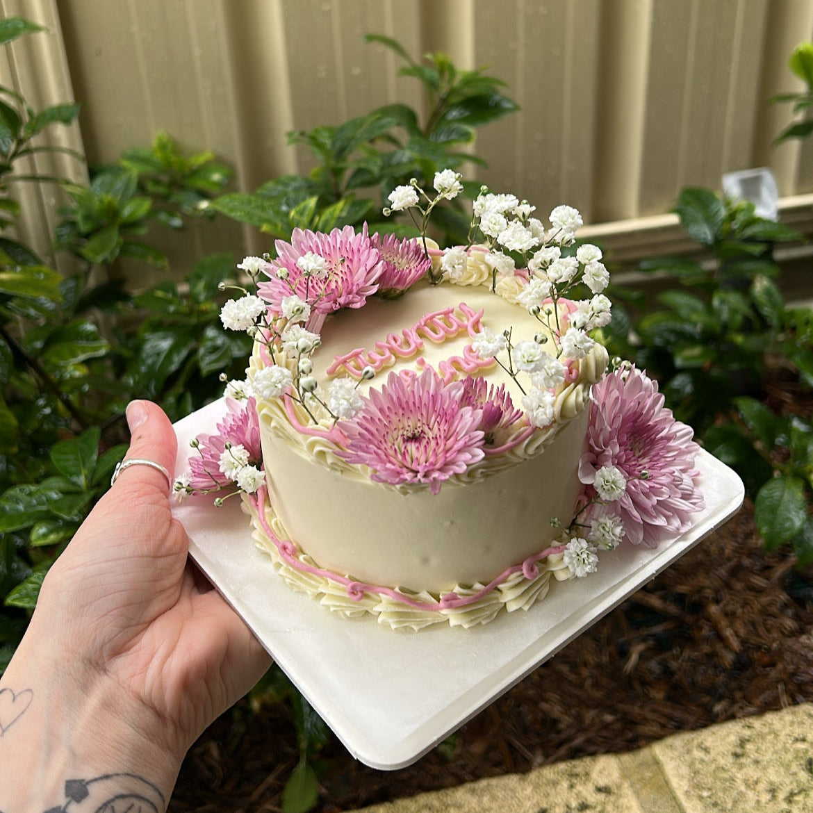 Small white bento cake with fresh pink and white flowers held by a hand against a green leafy background.