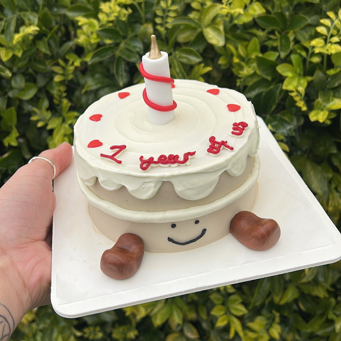 Top view of a small Jellycat inspired bento cake with a candle on top, and fondant Jellycat boots and anniversary writing on a white cake board being held in a hand, in front of a leafy green background.