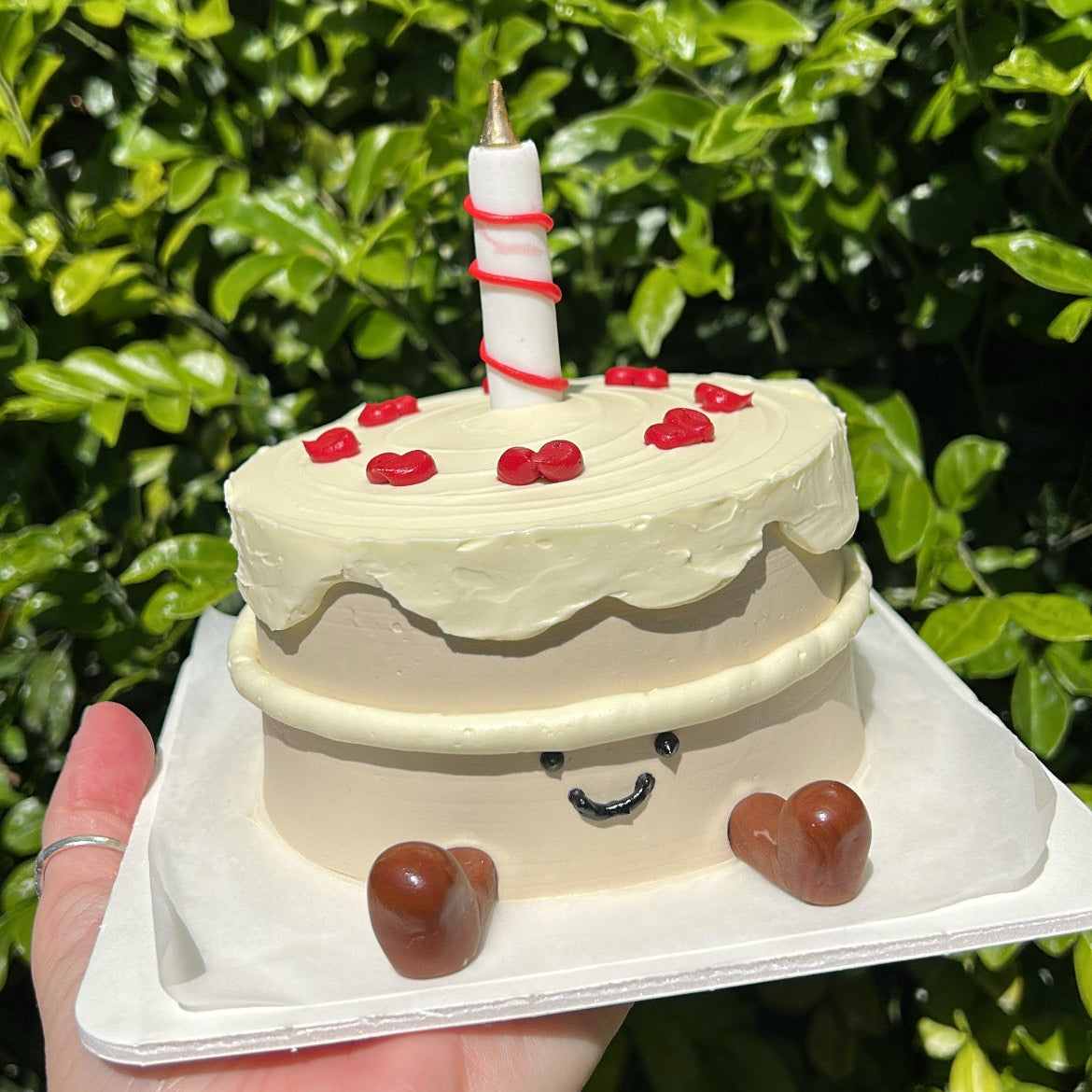 Side of a small Jellycat inspired bento cake with a candle on top, and fondant Jellycat boots on a white cake board being held in a hand, in front of a leafy green background.