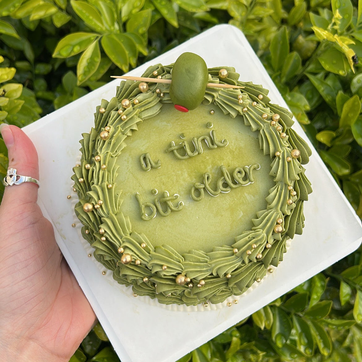 Top of a green cake with fondant olive and gold sprinkles, held by a hand against a green leafy background.