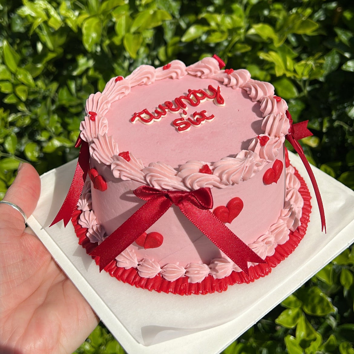 A small, round bento cake decorated with pink and red buttercream, red bows, and heart-shaped piping. The cake is held in a person's hand with a leafy green background.