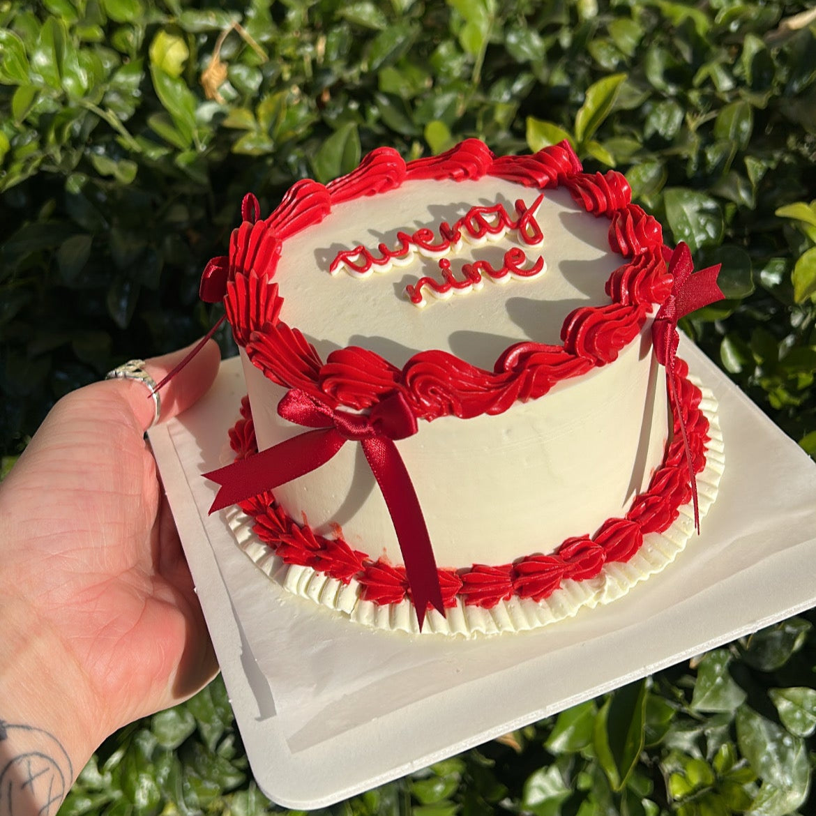 White bento cake with red frosting border, hand piped "twenty nine" and red rubbon bows, held by a hand against a leafy green background.