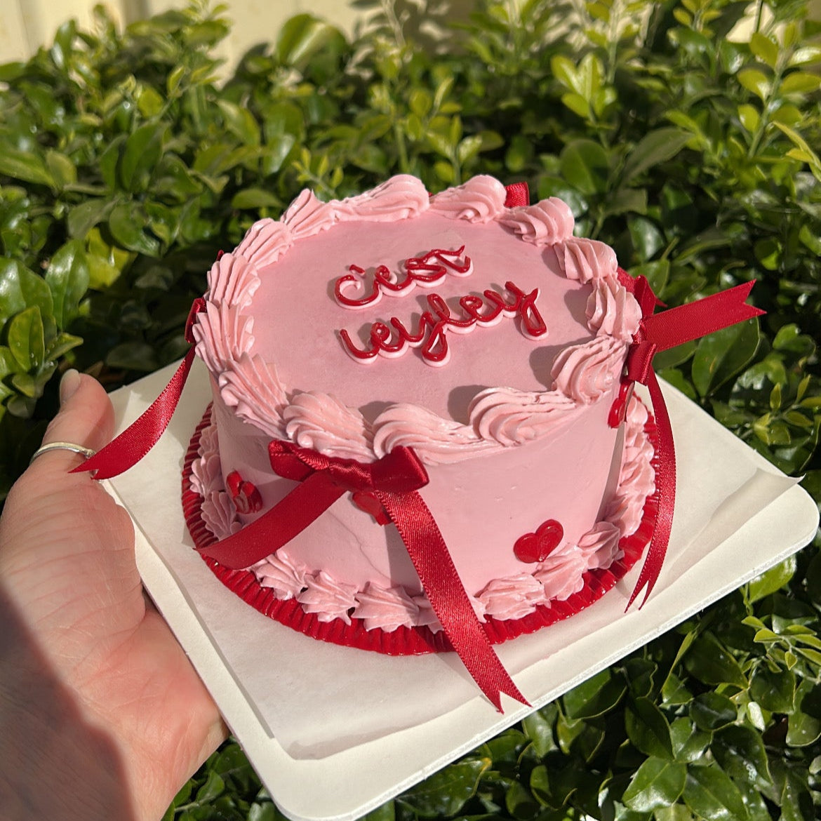 Pink cake with red ribbon held by a hand against a green bush background.