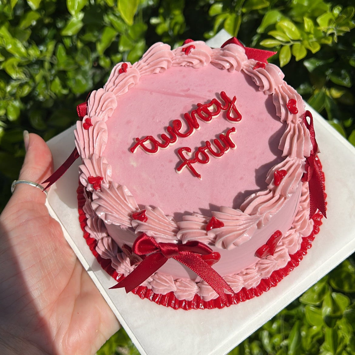 Top of a small pink and red bento cake, with red ribbon bows held by a hand against a leafy green background.