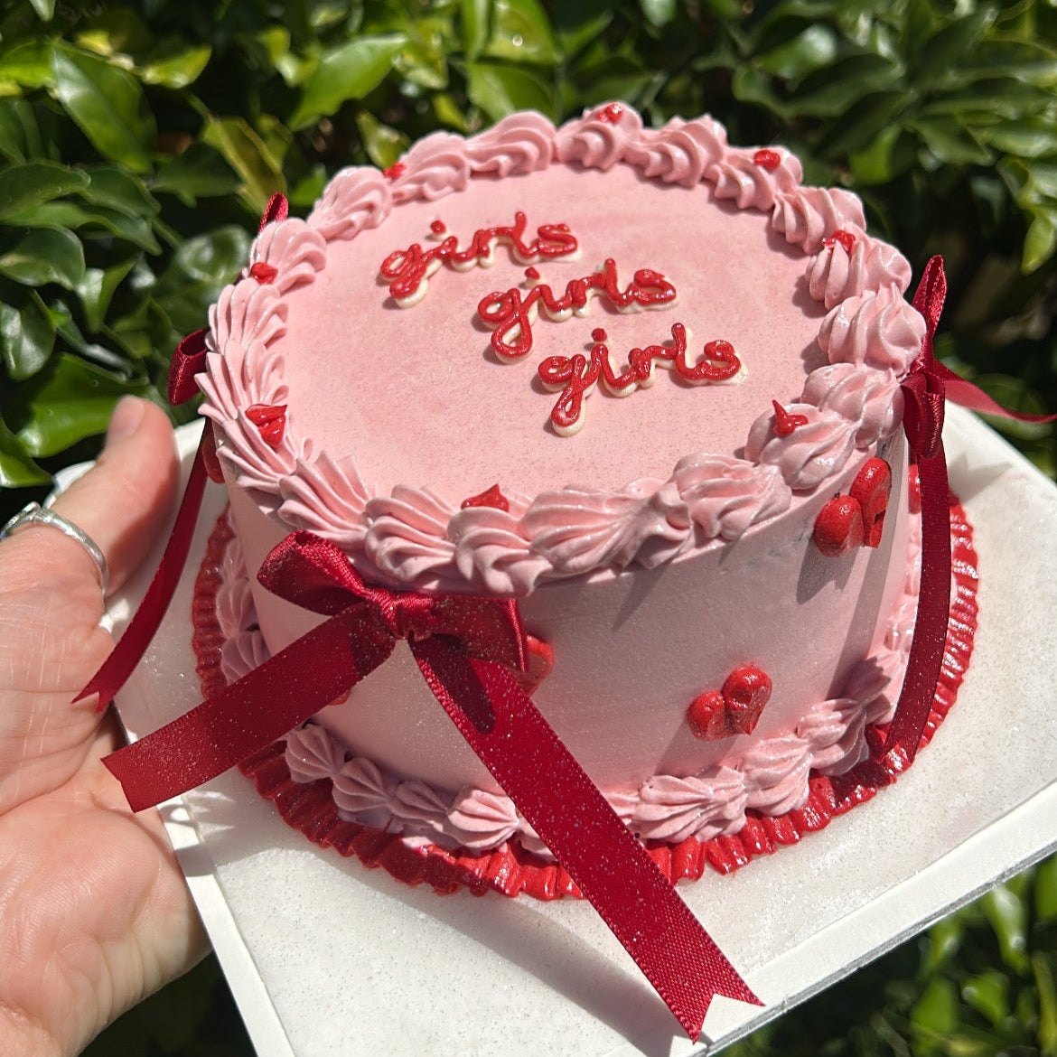 A bento cake with pink and red Swiss meringue buttercream frosting, decorated with red bows and the words 'girls girls girls' written on top.