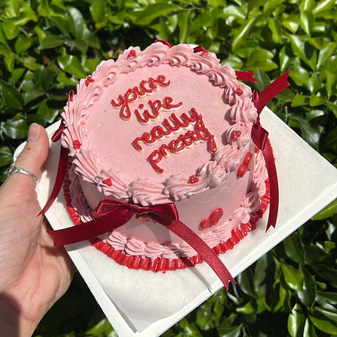 Top view of a pink bento cake, red heart and hand piping on top, with red bows, held by a hand against a green leafy background.