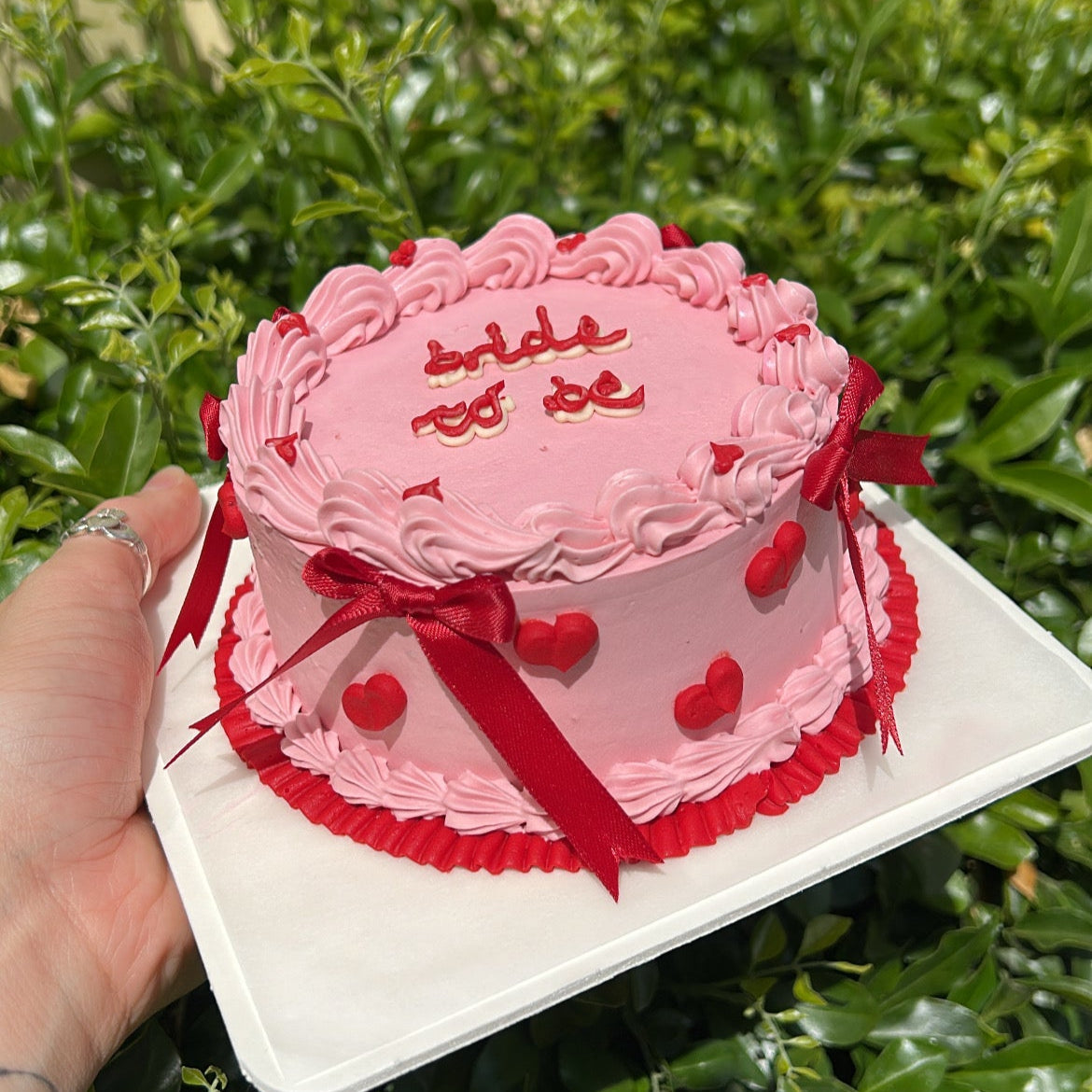 Side view of a pink bento cake with red heart piping and red ribbon bows, held against a leafy green background.