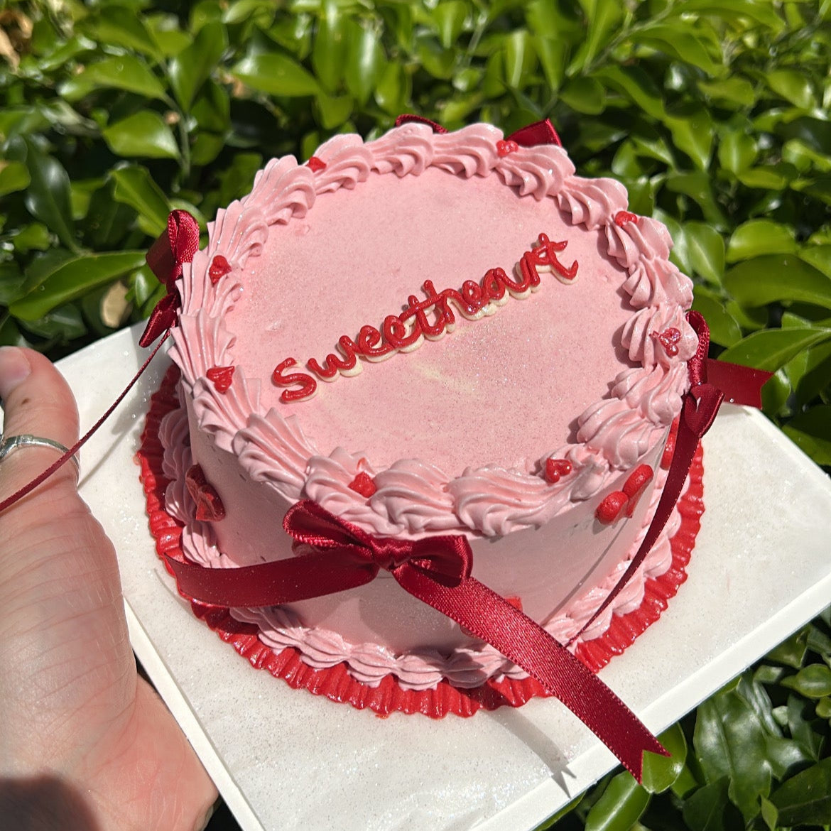 Top view of a pink "sweetheart" piped bento cake, with red decals and red bows. Held against a green leafy background.