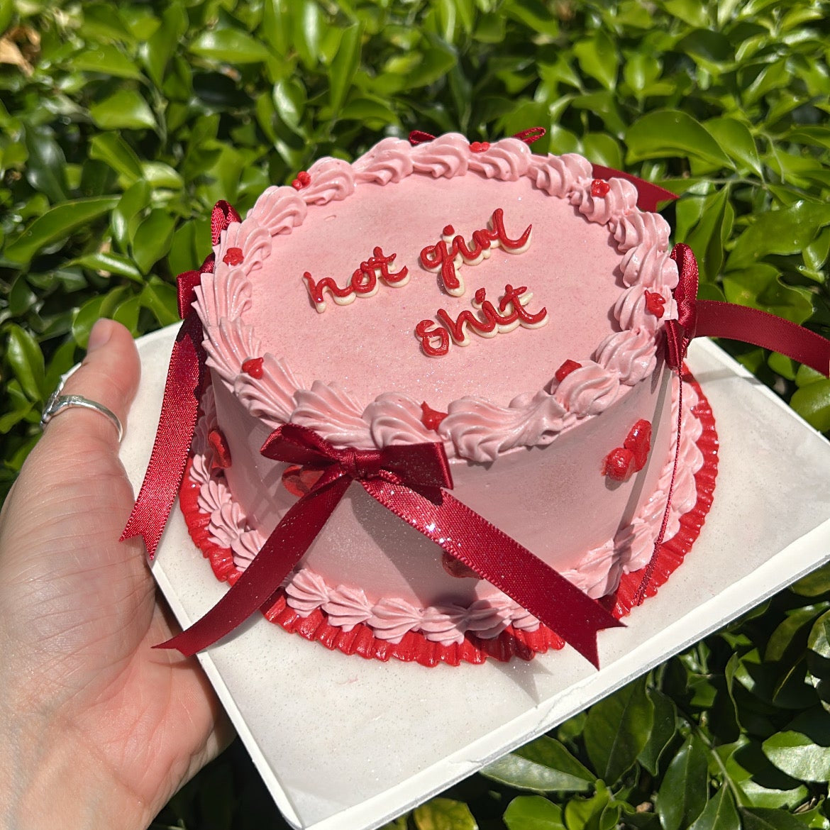 Pink and red small cake with red piping and red ribbon decals held by a hand against a green background.