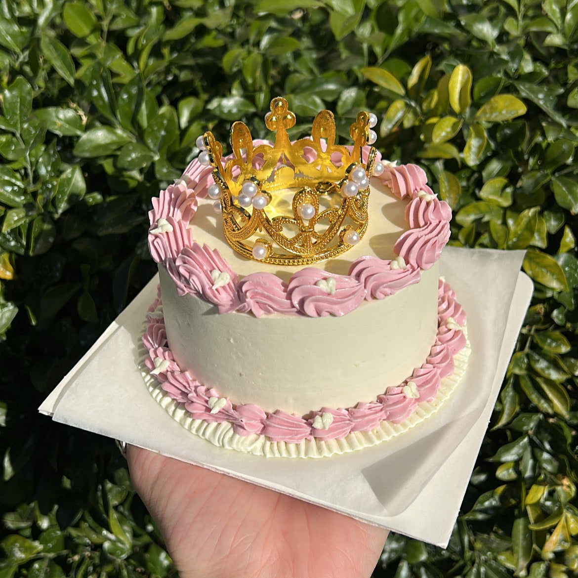 Side of a small white bento cake decorated with a pink buttercream border, topped with a golden crown, being held by a hand against a leafy background.
