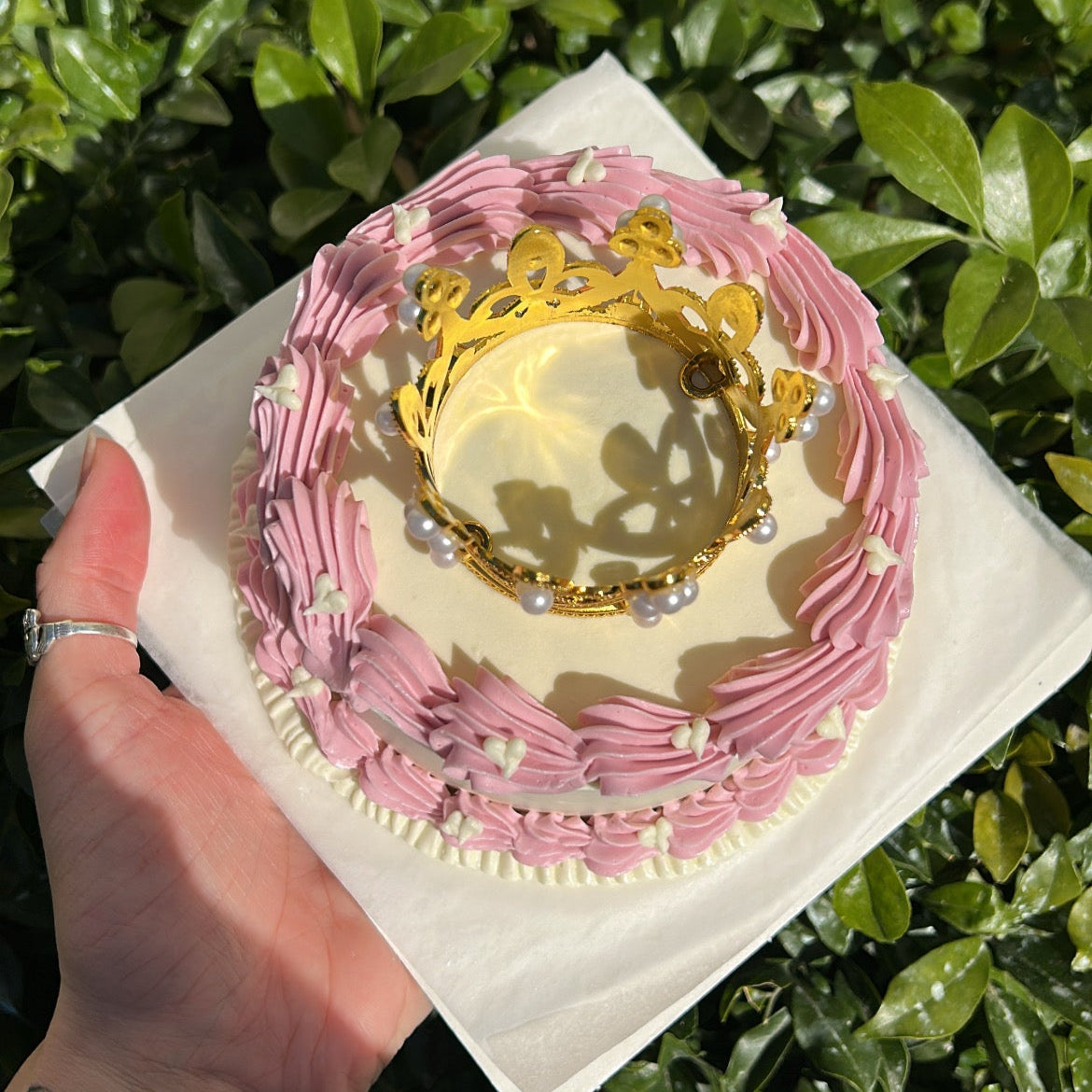 Top of a small white bento cake decorated with a pink buttercream border, topped with a golden crown, being held by a hand against a leafy background.