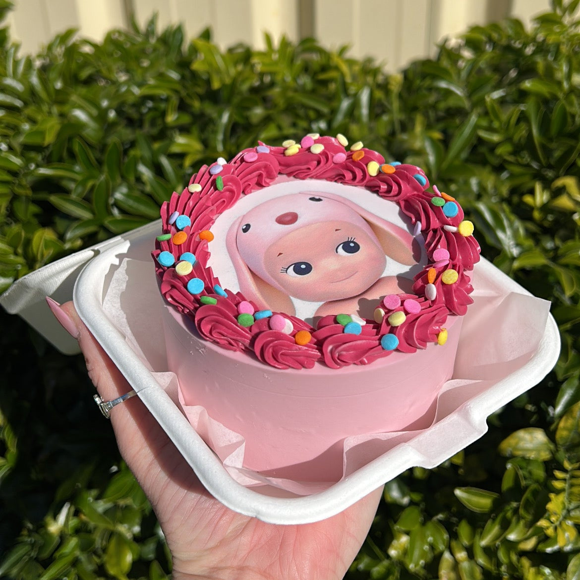 Small pink cake with pink bunny Sonny Angel edible image design, held in a hand against a green leafy background.