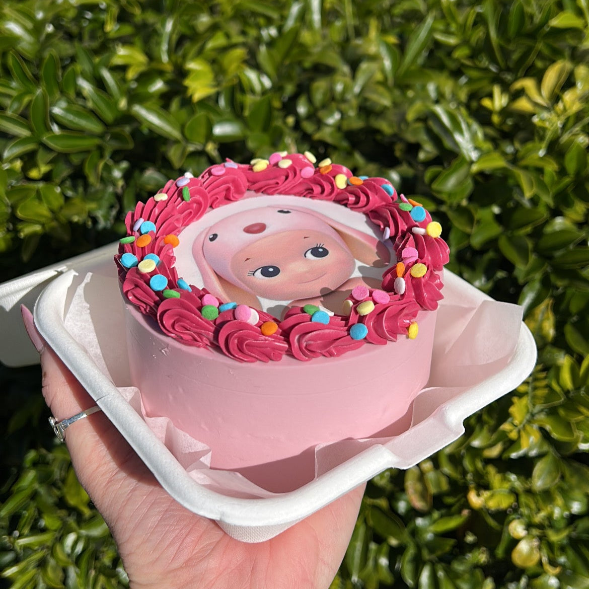 Side of a small pink cake with pink bunny Sonny Angel edible image design, held in a hand against a green leafy background.