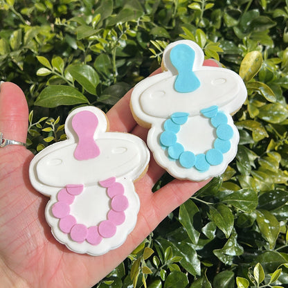 Pink and blue baby dummy sugar cookies held by a hand in front of a green leafy background.