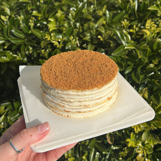 Layered Biscoff crepe cake with a Biscoff crumb topping, held on a white cake board against a green leafy background.