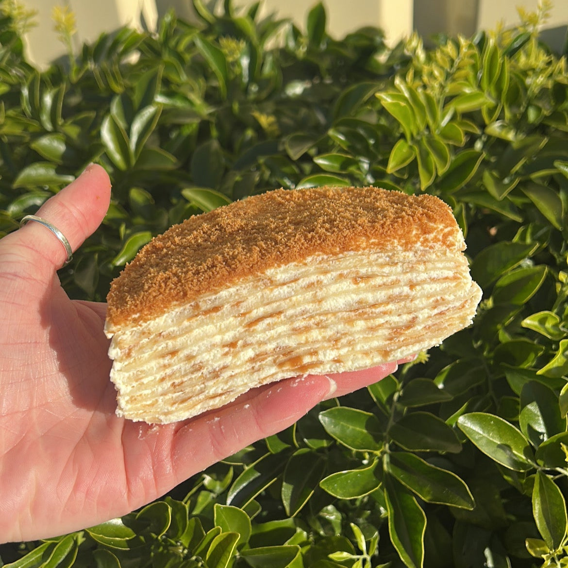 Inside of a layered Biscoff crepe cake with a Biscoff crumb topping, held on a white cake board against a green leafy background.