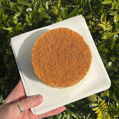 Top of a layered Biscoff crepe cake with a Biscoff crumb topping, held on a white cake board against a green leafy background.