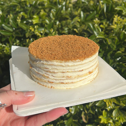 Side of a layered Biscoff crepe cake with a Biscoff crumb topping, held on a white cake board against a green leafy background.