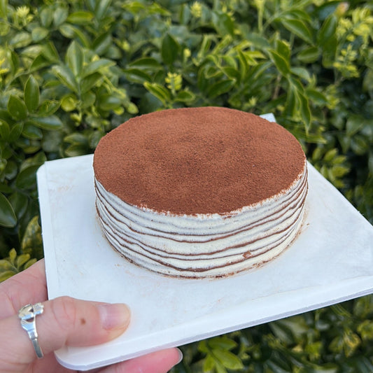 Layered chocolate crepe cake with a cocoa dusting topping, held on a white cake board against a green leafy background.