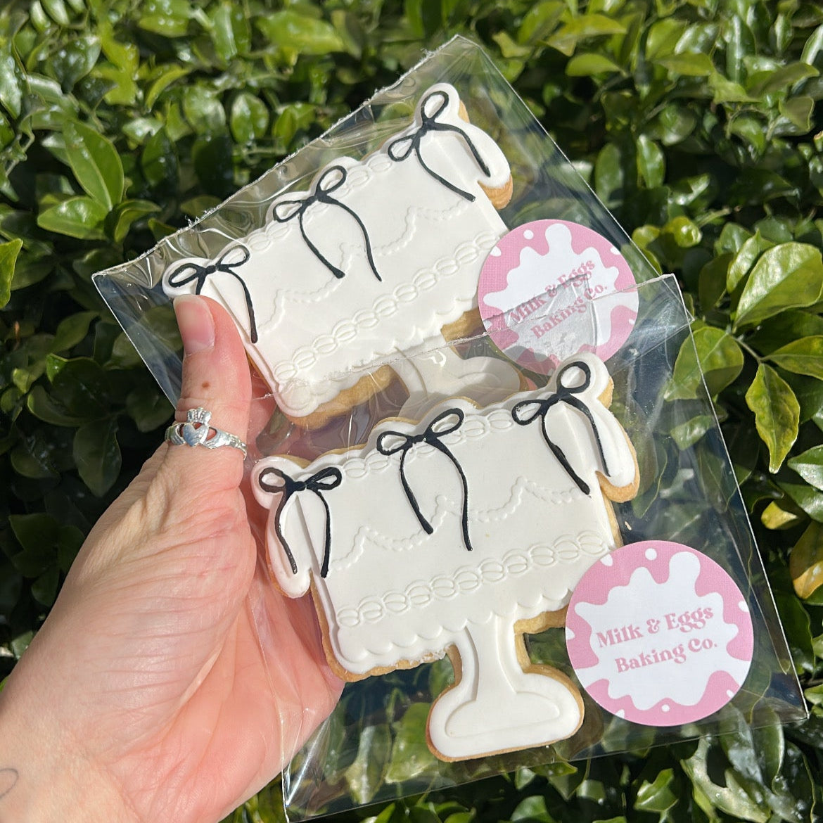 Packaged cookies shaped like cake with black coloured bows held by a hand in front of a green leafy background.