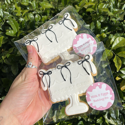 Packaged cookies shaped like cake with black coloured bows held by a hand in front of a green leafy background.