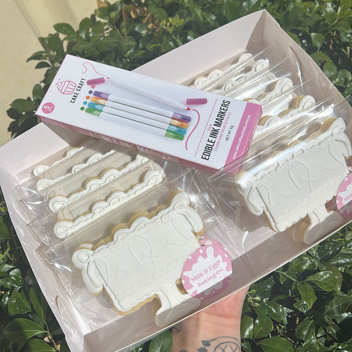 Packaged sugar cookies and a pack of edible markers with a hand holding them against a leafy green background.
