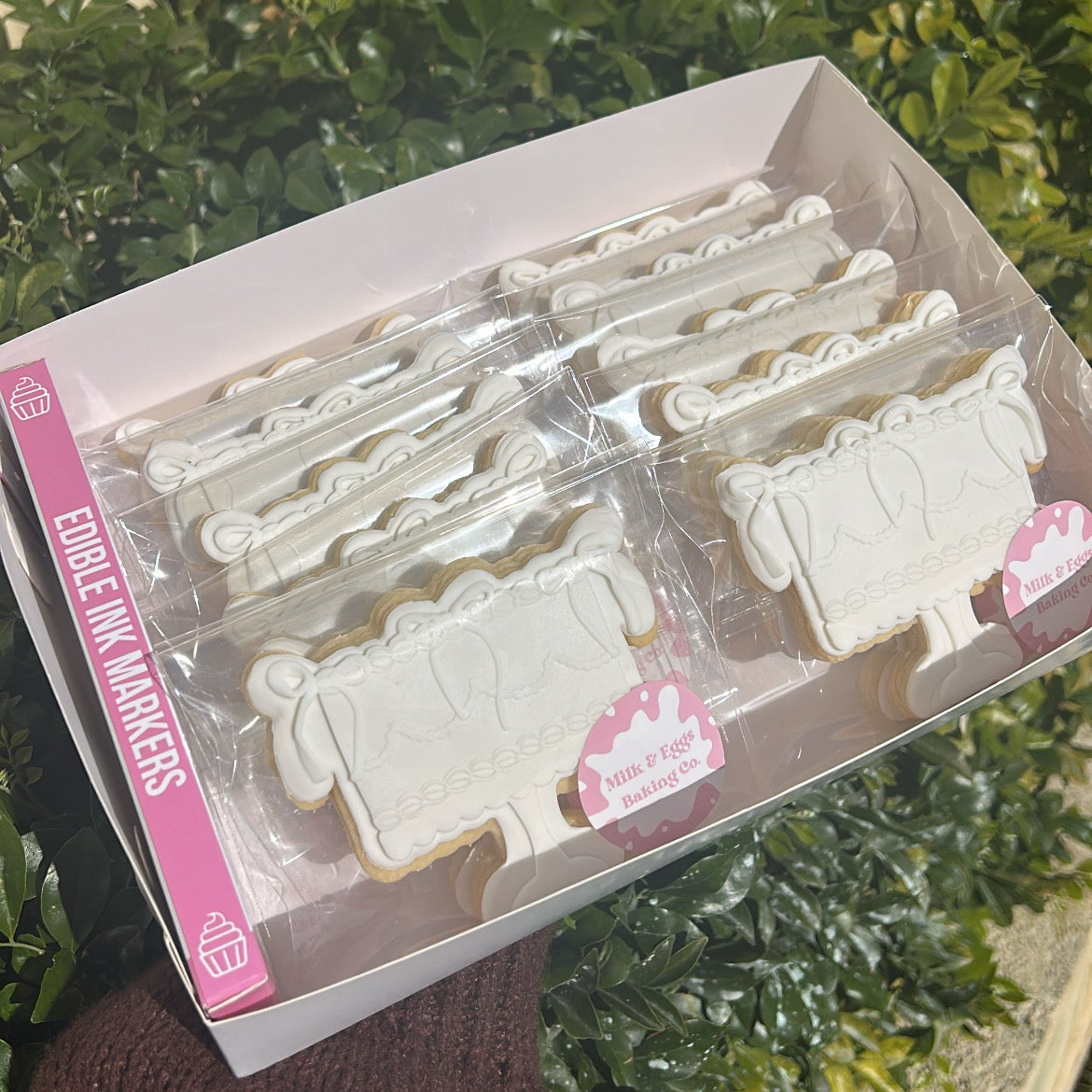 Packaged cake shaped sugar cookies with a pack of edible markers held in front of a leafy green background.