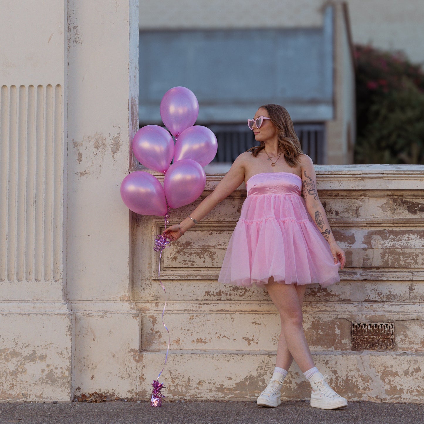 Woman in a pink dress holding pink balloons against a building backdrop.