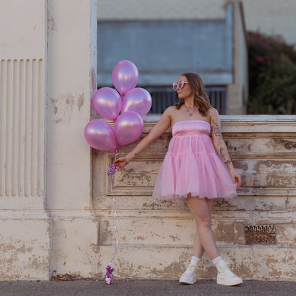 Woman in a pink dress holding pink balloons against a building backdrop.