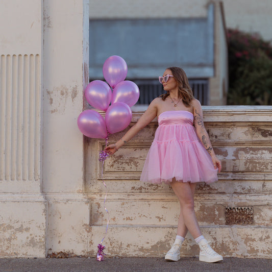 Woman in a pink dress holding pink balloons against a building backdrop.