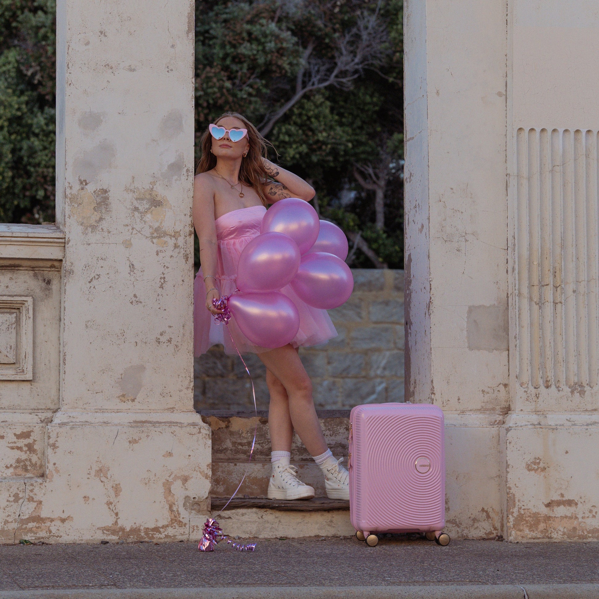 Woman in a pink dress with balloons and a pink suitcase standing in front of an archway.