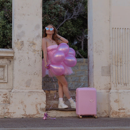 Woman in a pink dress with balloons and a pink suitcase standing in front of an archway.