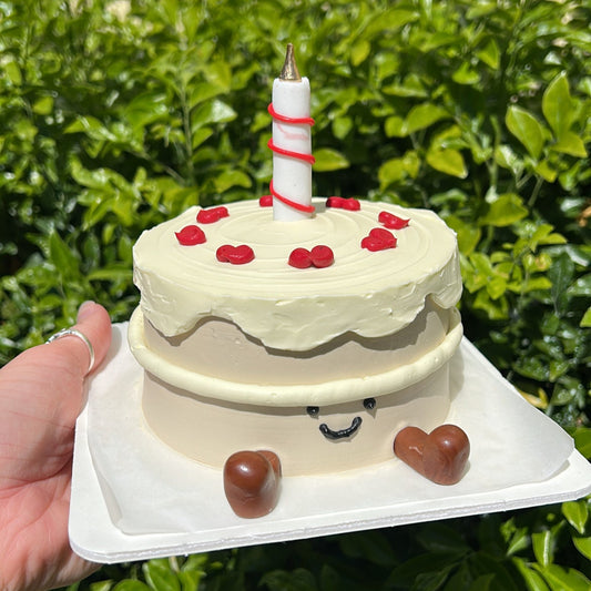 A small Jellycat inspired bento cake with a candle on top, and fondant Jellycat boots on a white cake board being held in a hand, in front of a leafy green background.