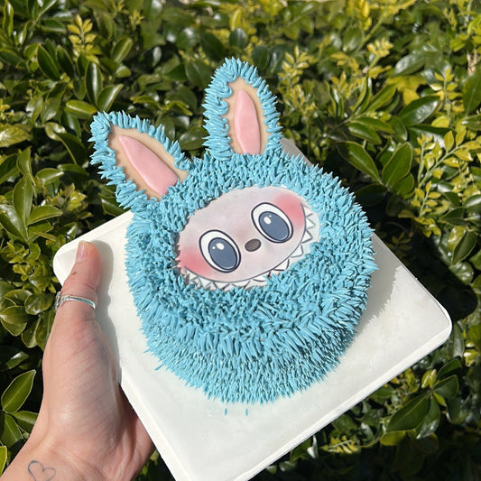 Small blue Labubu designed bento cake held in a hand against a green leafy background.