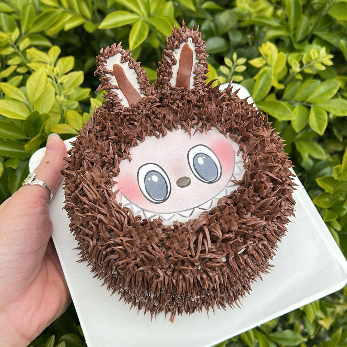 Small brown Labubu designed bento cake held in a hand against a green leafy background.