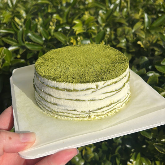 Layered matcha crepe cake with a matcha dusting topping, held on a white cake board against a green leafy background.