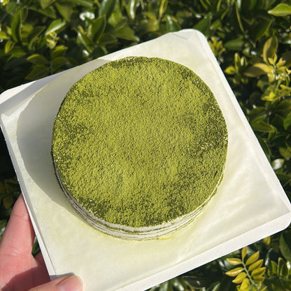 Top of a layered matcha crepe cake with a matcha dusting topping, held on a white cake board against a green leafy background.