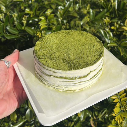 Side of a layered matcha crepe cake with a matcha dusting topping, held on a white cake board against a green leafy background.
