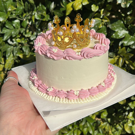 Small white bento cake decorated with a pink buttercream border, topped with a golden crown, being held by a hand against a leafy background.