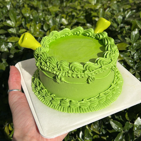 A small, lime green colored, Shrek inspired bento cake decorated with Swiss Meringue Buttercream frosting and topped with fondant Shrek ears, held in front of a leafy green background.