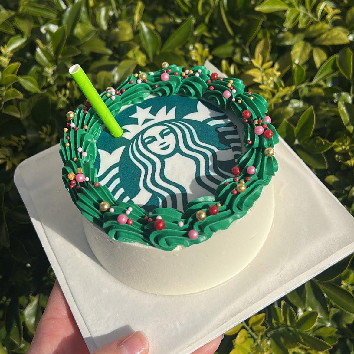 Starbucks-themed cake with green frosting, Starbucks logo edible image and a green straw on a white cake board against a green leafy background.
