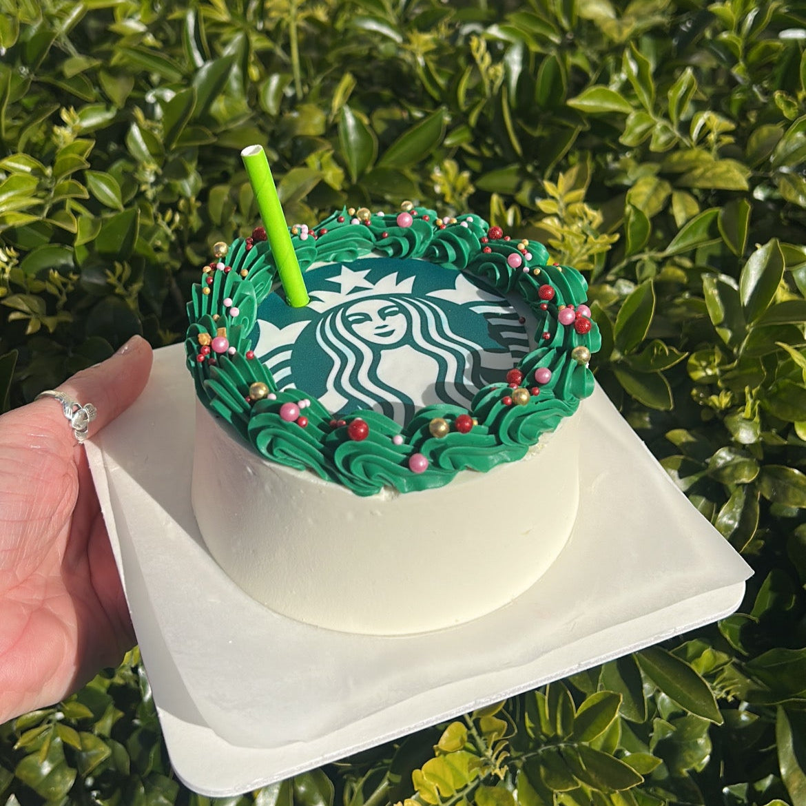 Side of a Starbucks-themed cake with green frosting, Starbucks logo edible image and a green straw on a white cake board against a green leafy background.