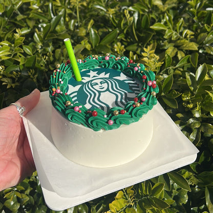 Side of a Starbucks-themed cake with green frosting, Starbucks logo edible image and a green straw on a white cake board against a green leafy background.