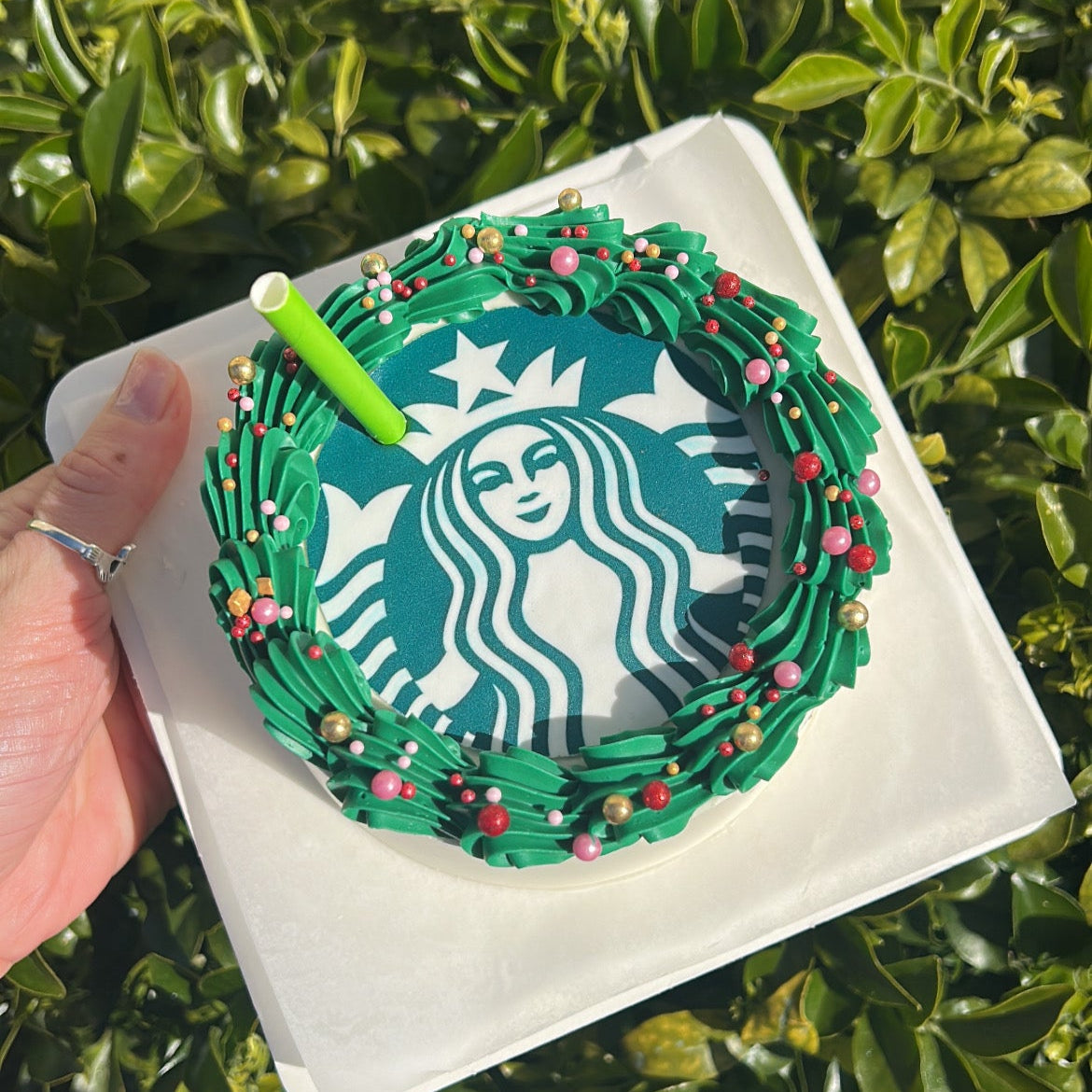 Top of a Starbucks-themed cake with green frosting, Starbucks logo edible image and a green straw on a white cake board against a green leafy background.