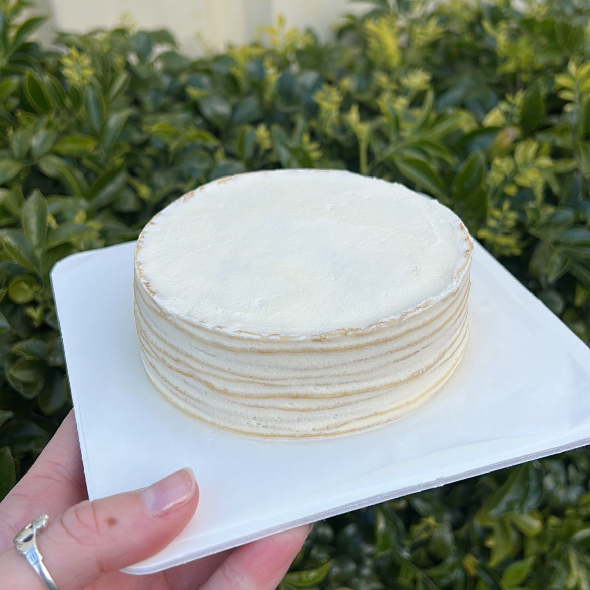Layered vanilla crepe cake with cream topping, held on a white cake board against a green leafy background.