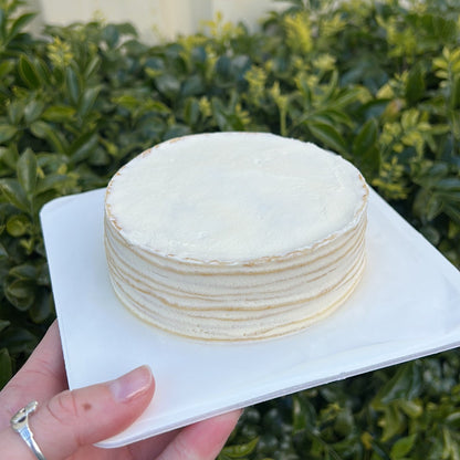 Layered vanilla crepe cake with cream topping, held on a white cake board against a green leafy background.