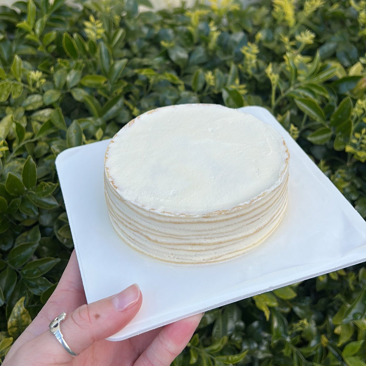 Side of a layered vanilla crepe cake with cream topping, held on a white cake board against a green leafy background.