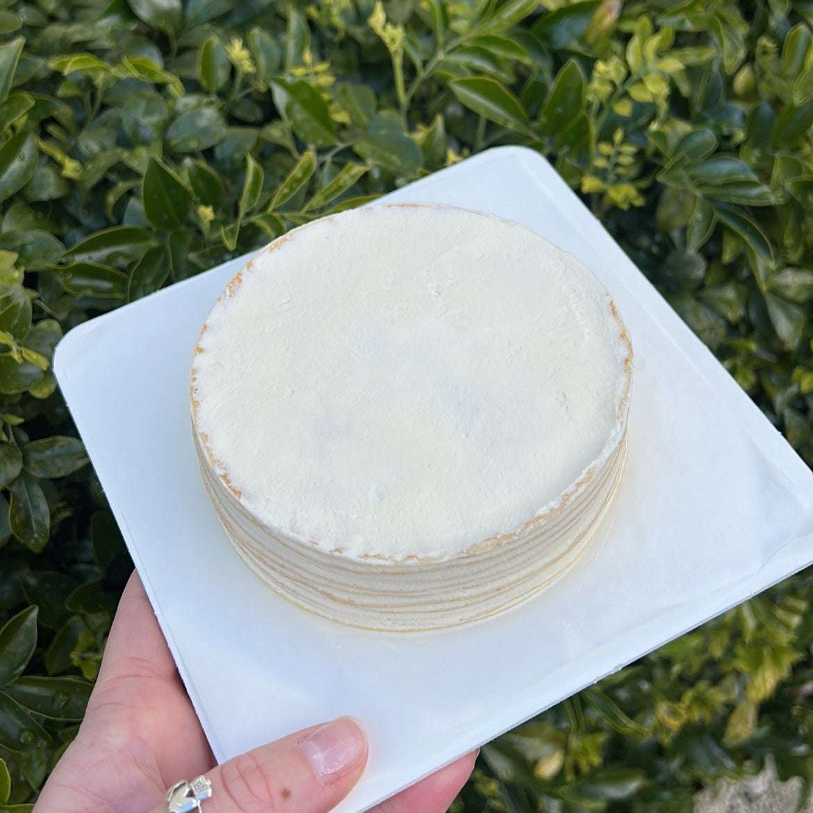 Top of a layered vanilla crepe cake with cream topping, held on a white cake board against a green leafy background.